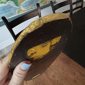 May include: A round, dark brown object with a yellow design and a gold-colored edge. The object is held in a person's hand. The object appears to be a decorative piece or part of a larger item.