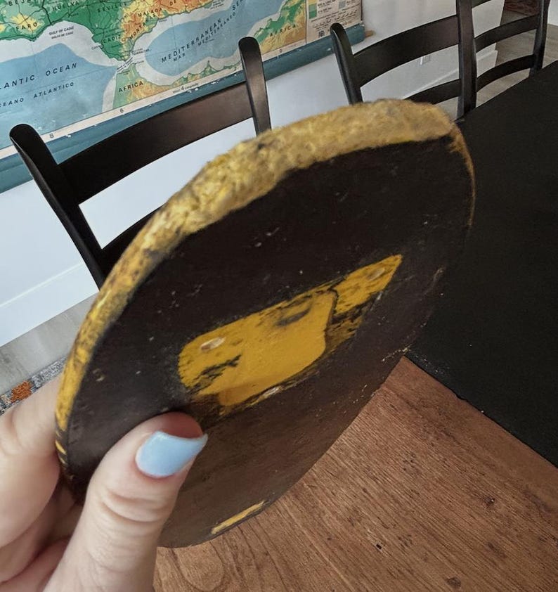 May include: A round, dark brown object with a yellow design and a gold-colored edge. The object is held in a person's hand. The object appears to be a decorative piece or part of a larger item.