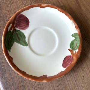 May include: A white ceramic saucer with a brown rim and fig illustrations. The saucer features a central recessed area for a cup and is decorated with green leaves and maroon figs.