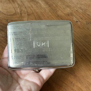 May include: Silver rectangular cigarette case with a textured surface and the initials "VK" engraved on the front. The case has a hinged lid and a polished finish, reflecting light. A vintage accessory.