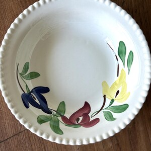 Op de afbeelding: Crèmekleurige keramische kom met een geschulpte rand. De kom is versierd met handgeschilderde bloemmotieven in blauw, geel en kastanjebruin, met groene bladeren. De kom staat op een houten oppervlak.