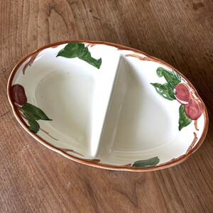 May include: Oval, divided serving dish with a cream-colored interior and a brown rim. The dish is decorated with green leaves and red-brown fruit. The dish is made of ceramic and is suitable for serving snacks or appetizers.