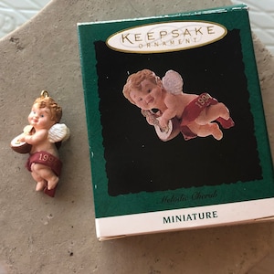 May include: A miniature porcelain cherub ornament playing a lute. The ornament is wearing a red sash with the year 1994 printed on it. The ornament is in its original box, which is green and white and says "Keepsake Ornament" and "Melodie Cherub Miniature".