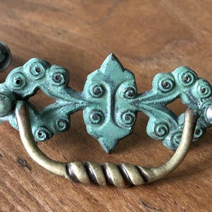 May include: Antique-style drawer pull with a distressed teal finish and a brass handle. The decorative hardware features an ornate design with swirling details and a twisted handle, perfect for adding a vintage touch to furniture.