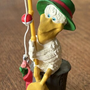 May include: A white bird with a yellow beak and blue eyes is wearing a green hat with a red band. The bird is holding a fishing rod with a red and white bobber. The bird is sitting on a gray log.