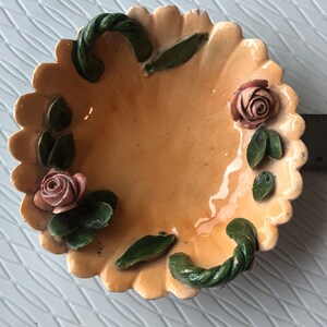 May include: A small, cream-colored ceramic dish with a scalloped edge. The dish is decorated with green leaves and pink roses. It has two green rope-like handles.