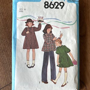 Puede incluir: Patrón de costura Simplicity vintage 8629 para ropa de niña. El patrón presenta ilustraciones de tres niñas con vestidos y pantalones. El paquete es azul y blanco con el logotipo de Simplicity e información de tallas.
