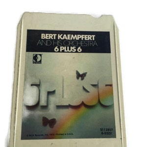 Peut inclure: Une cassette audio vintage avec un boîtier blanc et une image colorée d'un arc-en-ciel et de papillons. Le texte sur la cassette indique "Bert Kaempfert and His Orchestra 6 Plus 6".