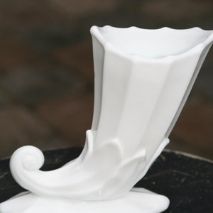 May include: A white ceramic cornucopia vase with a fluted design. The vase has a curved handle and a wide opening at the top.