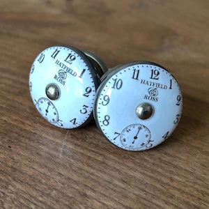 Puede incluir: Dos pomos de armario blancos con diseños de esfera de reloj, números negros y el texto "HATFIELD ROSS". Cada pomo tiene un centro plateado y una base plateada. Los pomos están sobre una superficie de madera.