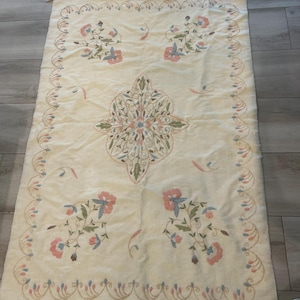 May include: A rectangular cream-colored textile featuring a central symmetrical floral medallion and four corner floral clusters. The border is decorated with a repeating vine pattern, and the short ends are finished with knotted fringe tassels.