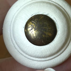 Peut inclure: Gros plan sur un bouton de meuble en bois peint en blanc avec un design circulaire. Le centre pr&eacute;sente un disque m&eacute;tallique de couleur laiton. Le bouton est probablement destin&eacute; aux meubles tels que les tiroirs ou les armoires.