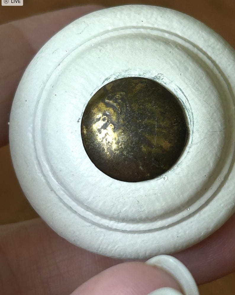 Peut inclure: Gros plan sur un bouton de meuble en bois peint en blanc avec un design circulaire. Le centre pr&eacute;sente un disque m&eacute;tallique de couleur laiton. Le bouton est probablement destin&eacute; aux meubles tels que les tiroirs ou les armoires.