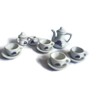 May include: A miniature white porcelain tea set with a teapot, sugar bowl, creamer, and several teacups with saucers. Each piece is decorated with a purple rose design. The set is ideal for a dollhouse or a child's play set.