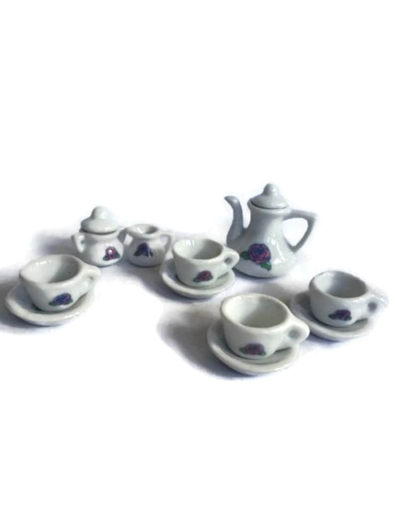May include: A miniature white porcelain tea set with a teapot, sugar bowl, creamer, and several teacups with saucers. Each piece is decorated with a purple rose design. The set is ideal for a dollhouse or a child's play set.