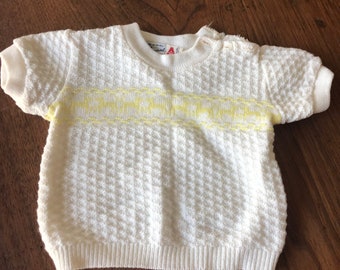 May include: White knitted baby sweater with short sleeves and a yellow stripe across the chest. The sweater has a waffle knit texture.