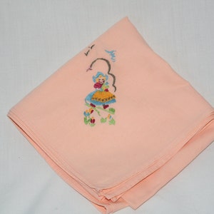 May include: A peach-colored fabric handkerchief with an embroidered design of a girl in a blue dress and yellow skirt, surrounded by flowers.