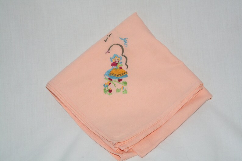 May include: A peach-colored fabric handkerchief with an embroidered design of a girl in a blue dress and yellow skirt, surrounded by flowers.