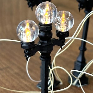 Pode incluir: Miniaturas de postes de luz pretos com luzes de globo transparentes e brilhantes. As lâmpadas têm um design clássico com um poste central e braços decorativos que seguram as luzes. Fios brancos são visíveis.