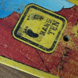 May include: Close-up of a vintage skateboard deck with a distressed finish. The deck features a yellow square with the text "HANG TEN" and a footprint graphic. The background has red and blue paint with visible wear and tear.