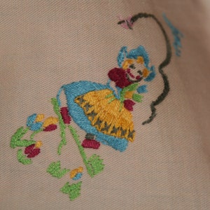May include: Embroidered design of a girl in a blue and yellow dress holding a flower. The design is stitched on a light pink fabric.