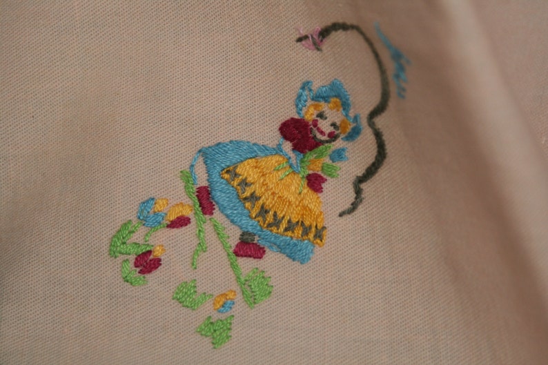 May include: Embroidered design of a girl in a blue and yellow dress holding a flower. The design is stitched on a light pink fabric.