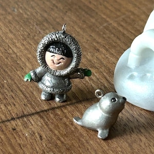 以下が含まれることがあります： 笑顔の人物がフード付きのパーカーを着て、もう一方はアザラシを描いた、2つのシルバーチャームペンダント。両方のチャームには、吊り下げ用のループが付いています。