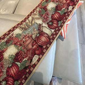May include: A rectangular tapestry table runner featuring a fall harvest theme. The runner displays pumpkins, apples, and floral arrangements in shades of red, orange, and green. The border has a red and brown checkered pattern.