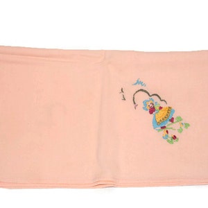 May include: A pale pink fabric square with a small embroidered design of a girl in a blue dress and yellow skirt. The girl is surrounded by green leaves and pink flowers.