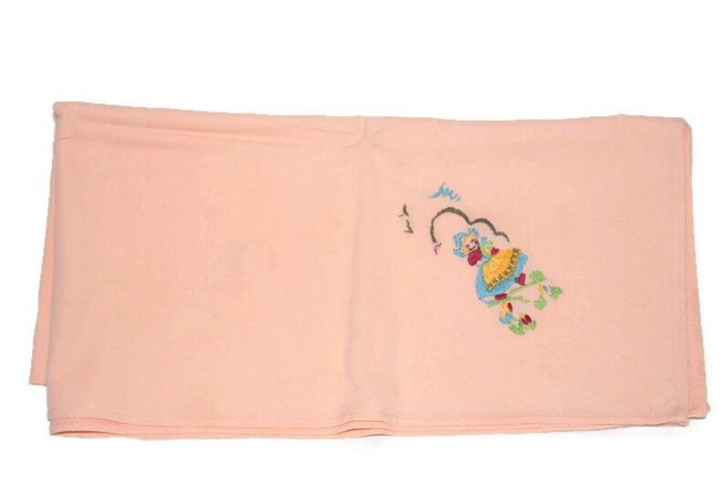 May include: A pale pink fabric square with a small embroidered design of a girl in a blue dress and yellow skirt. The girl is surrounded by green leaves and pink flowers.