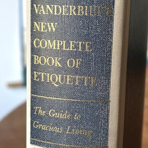 May include: A close-up of a book spine with the title "Amy Vanderbilt's New Complete Book of Etiquette." The cover is a textured navy blue with gold lettering. The book is a guide to gracious living, published by Doubleday.