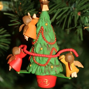 May include: A Christmas tree ornament featuring a green Christmas tree with red beads, a gold star on top, and three angels hanging from the branches. The angels are wearing orange, red, and yellow robes.