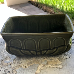 May include: A rectangular, olive green ceramic planter with a raised, arched design around the base. The planter has a glossy finish and sits on small feet. The interior is empty, ready for plants or flowers.