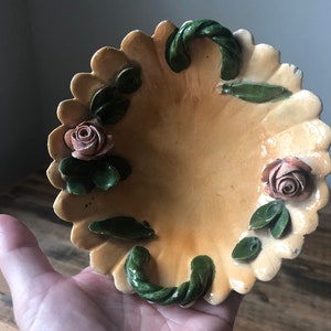 May include: A small, oval-shaped ceramic dish with a scalloped edge. The dish is decorated with green leaves and pink roses in a raised design. The dish has two green rope-like handles.