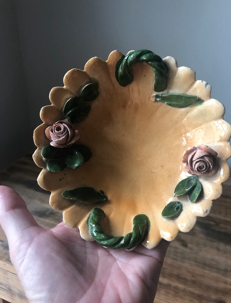 May include: A small, oval-shaped ceramic dish with a scalloped edge. The dish is decorated with green leaves and pink roses in a raised design. The dish has two green rope-like handles.