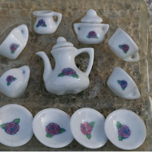 May include: A miniature white porcelain tea set with a floral rose design. The set includes a teapot, sugar bowl, creamer, cups, and saucers. Each piece features a red and blue rose with green leaves.