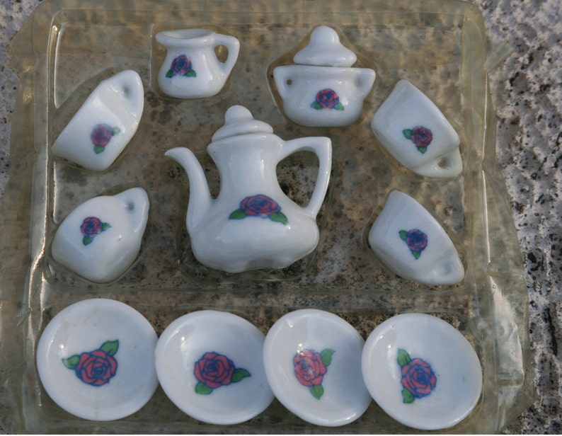 May include: A miniature white porcelain tea set with a floral rose design. The set includes a teapot, sugar bowl, creamer, cups, and saucers. Each piece features a red and blue rose with green leaves.