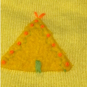 May include: A yellow felt appliqu&eacute; of a teepee with orange stitching and green details. The teepee is on a yellow knitted fabric.