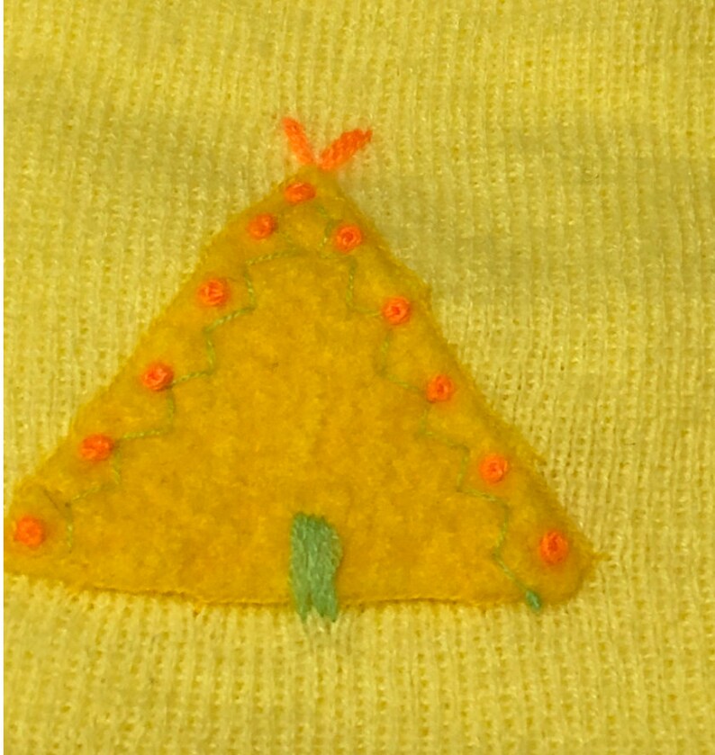 May include: A yellow felt appliqu&eacute; of a teepee with orange stitching and green details. The teepee is on a yellow knitted fabric.