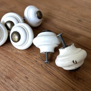 Peut inclure: Ensemble de boutons de meubles blancs, &agrave; finition vieillie, avec des ferrures couleur laiton. Les boutons sont ronds avec un design d&eacute;coratif, pr&eacute;sent&eacute;s sur une surface en bois. Certains boutons ont des vis.