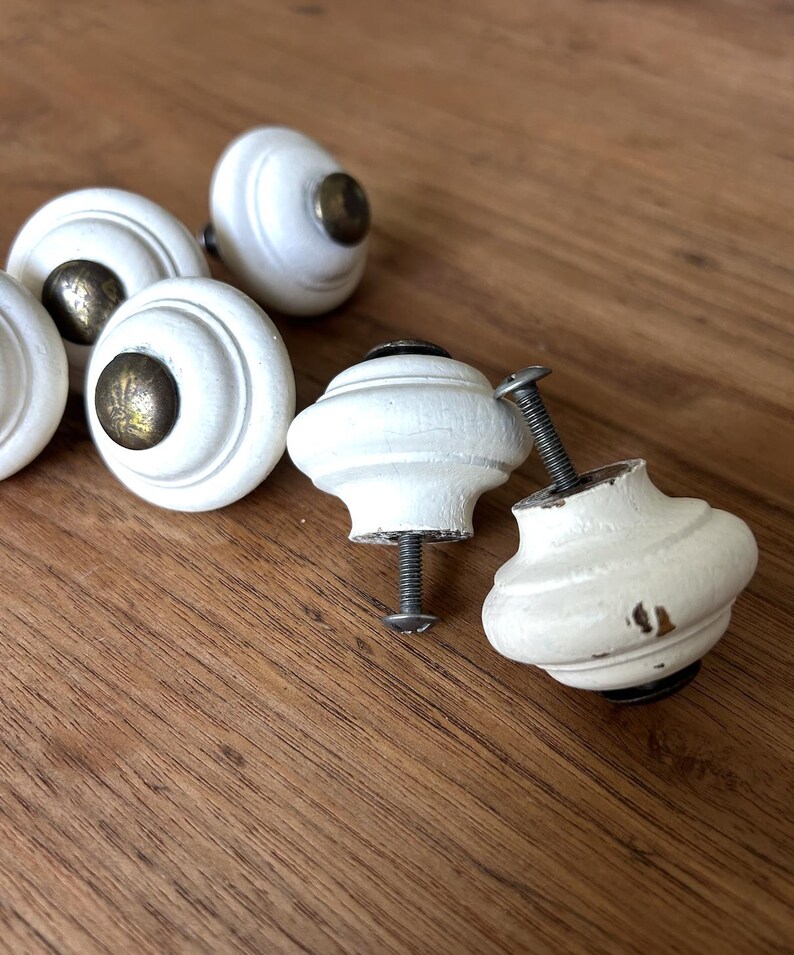 Peut inclure: Ensemble de boutons de meubles blancs, &agrave; finition vieillie, avec des ferrures couleur laiton. Les boutons sont ronds avec un design d&eacute;coratif, pr&eacute;sent&eacute;s sur une surface en bois. Certains boutons ont des vis.