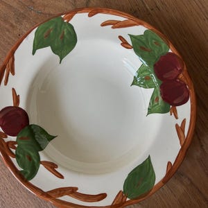 May include: A white ceramic serving bowl with a decorative rim and painted design. The rim is brown with a branch-like pattern. Green leaves and maroon fruit are painted around the bowl's edge. A classic piece for serving salads or pasta.