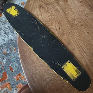 May include: A vintage skateboard with a black deck and yellow accents. The deck shows wear and tear, with yellow mounting plates at each end. The board is set against a wooden surface.