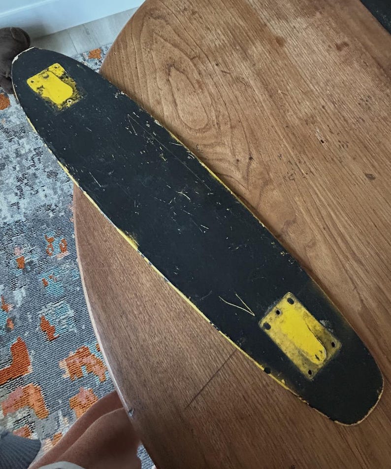 May include: A vintage skateboard with a black deck and yellow accents. The deck shows wear and tear, with yellow mounting plates at each end. The board is set against a wooden surface.