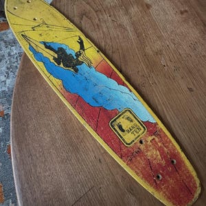 May include: Vintage skateboard with a yellow top, featuring a graphic of a person surfing a blue wave against a red and yellow background. The board has the words "Hang Ten" on a rectangular label.