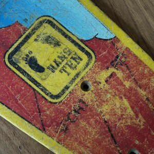 May include: Close-up of a vintage skateboard deck with a distressed finish. The deck features a yellow and red color scheme with a black and yellow sign that reads "Hang Ten" and a footprint graphic. Two holes are visible.