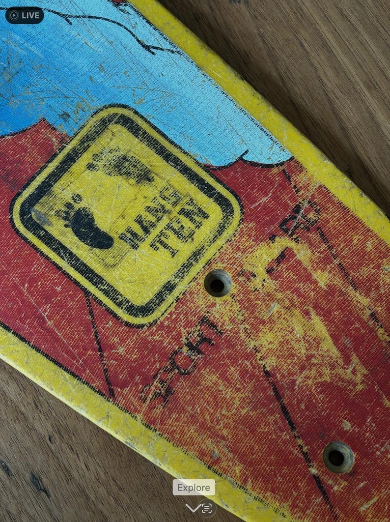 May include: Close-up of a vintage skateboard deck with a distressed finish. The deck features a yellow and red color scheme with a black and yellow sign that reads "Hang Ten" and a footprint graphic. Two holes are visible.