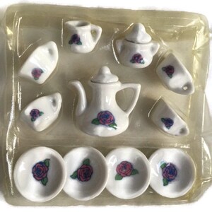 May include: A miniature white porcelain tea set with a teapot, cups, saucers, and a sugar bowl. Each piece is decorated with a red and blue rose design. The set is packaged in a clear plastic container.