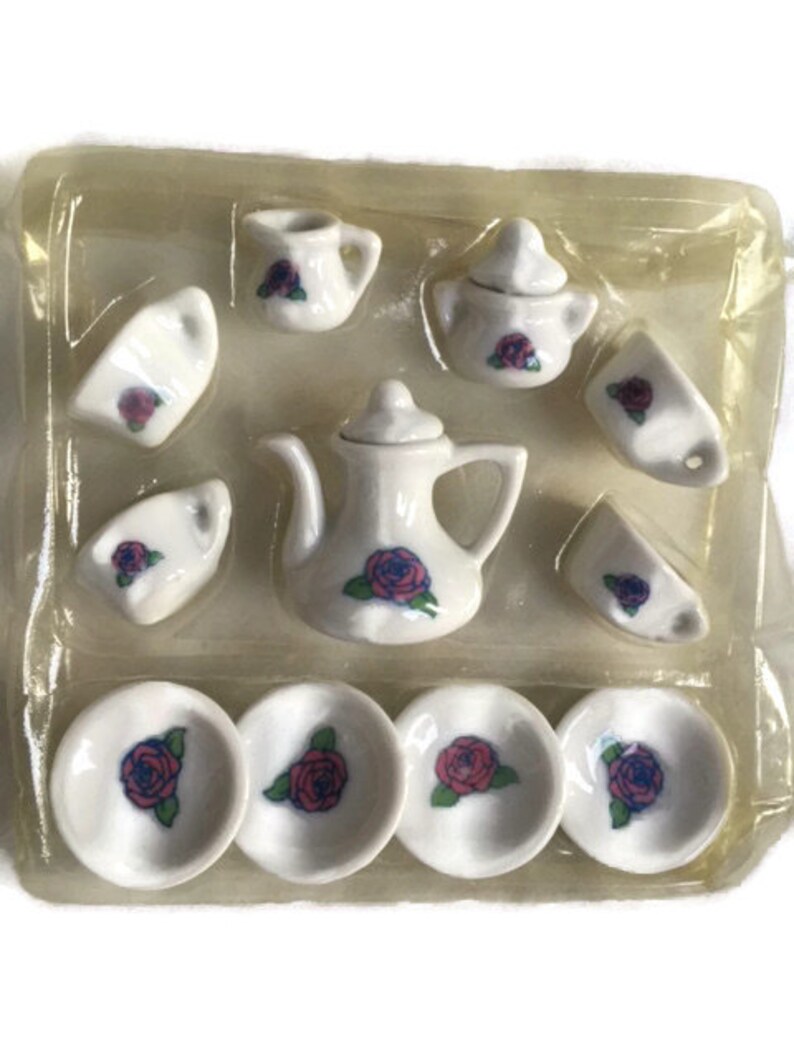 May include: A miniature white porcelain tea set with a teapot, cups, saucers, and a sugar bowl. Each piece is decorated with a red and blue rose design. The set is packaged in a clear plastic container.