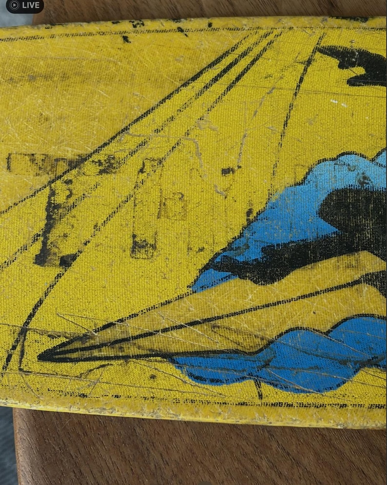 May include: Close-up of a yellow, textured surface with a vintage illustration. The artwork features black lines radiating from the top and blue cloud-like shapes. The piece has a distressed appearance, suggesting age and wear.